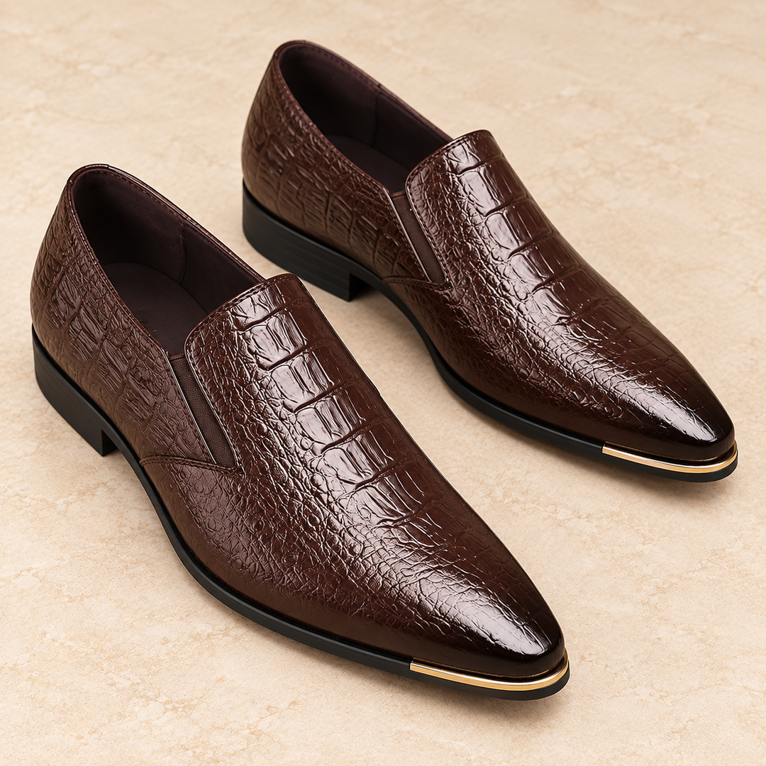Close three-quarter studio view of Callum dark brown pointed loafers showing the embossed vamp, gold-tone toe trim and black sole.