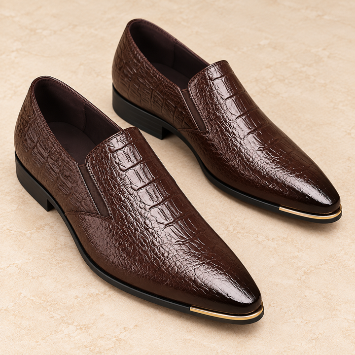 Close three-quarter studio view of Callum dark brown pointed loafers showing the embossed vamp, gold-tone toe trim and black sole.