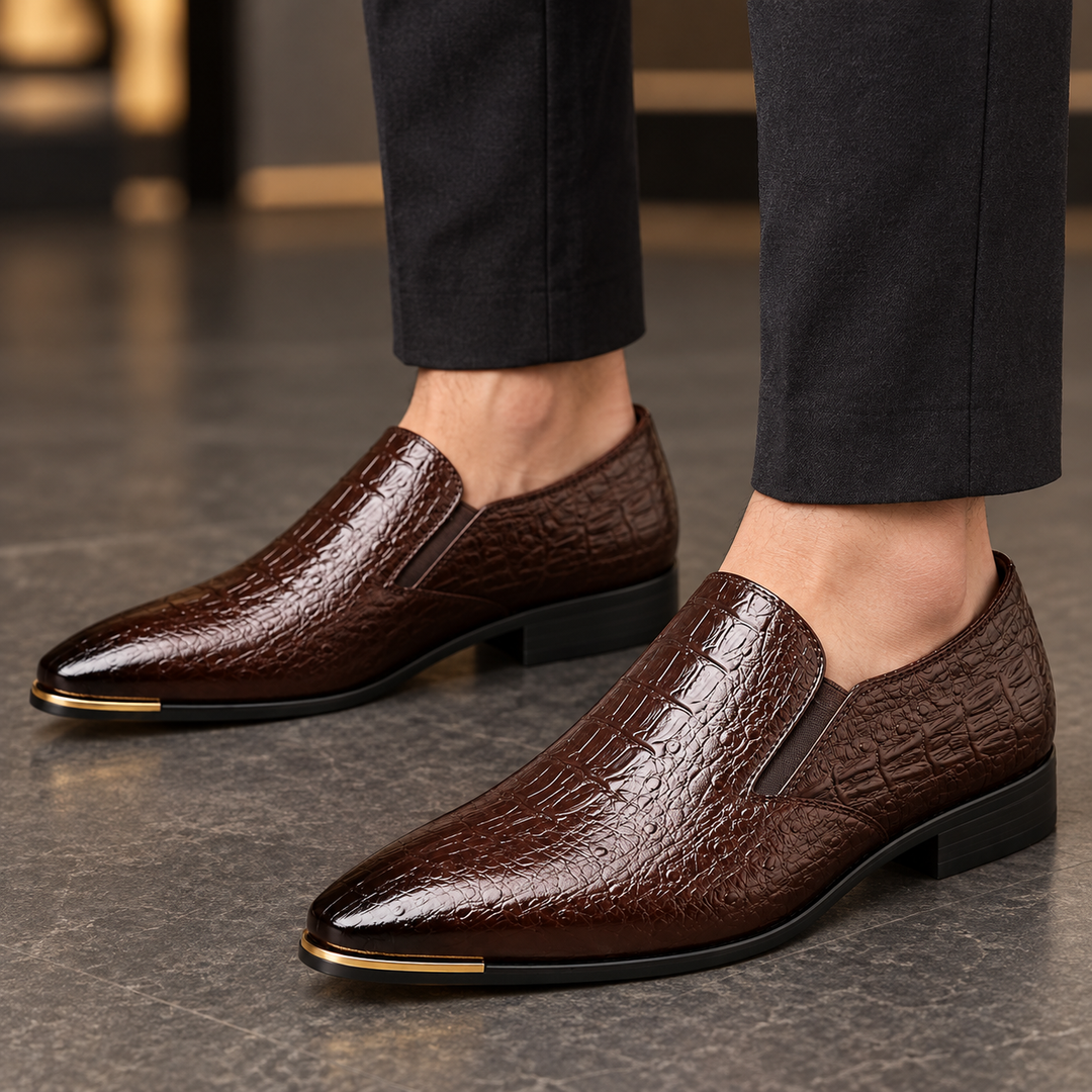 Callum dark brown crocodile-embossed loafers worn with tailored trousers, framed closely around the shoes and ankles.