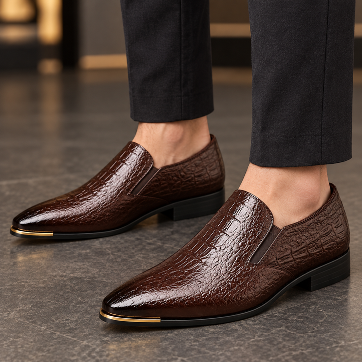 Callum dark brown crocodile-embossed loafers worn with tailored trousers, framed closely around the shoes and ankles.